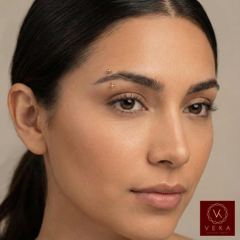 Close-up of a woman's face with real 14k gold Eyebrow piercing with a neutral background and Veka logo.