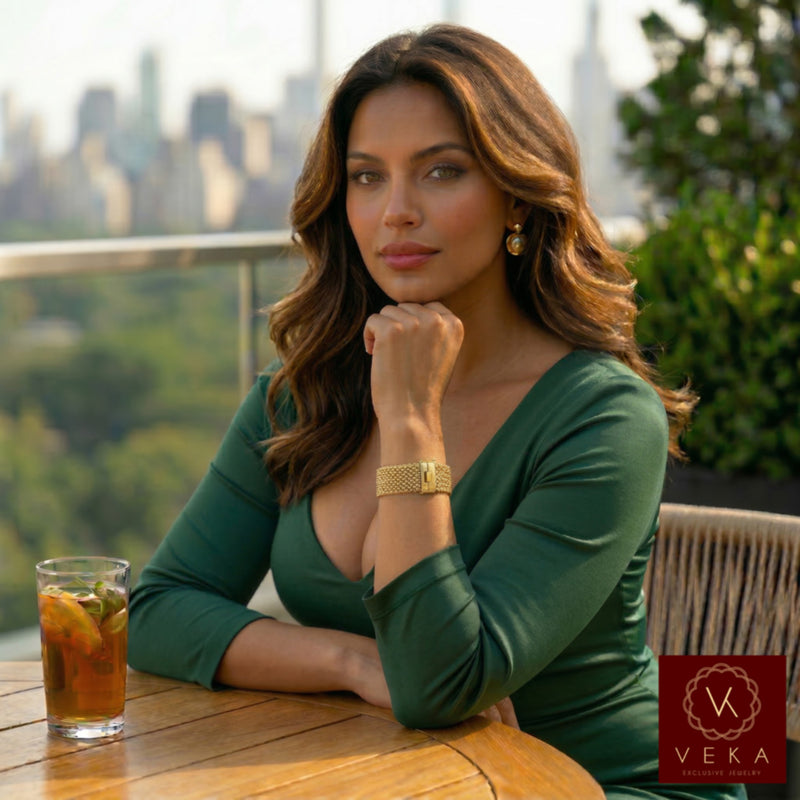 A high-resolution, professional photo of a model with long wavy brown hair, wearing a dark green V-neck dress. She is seated at a wooden table on a city terrace with a blurred skyline background. Her hand is resting under her chin, prominently displaying a thick, shimmering 14k gold popcorn-link bracelet on her wrist. The lighting is warm and natural, highlighting the detailed texture of the jewelry.