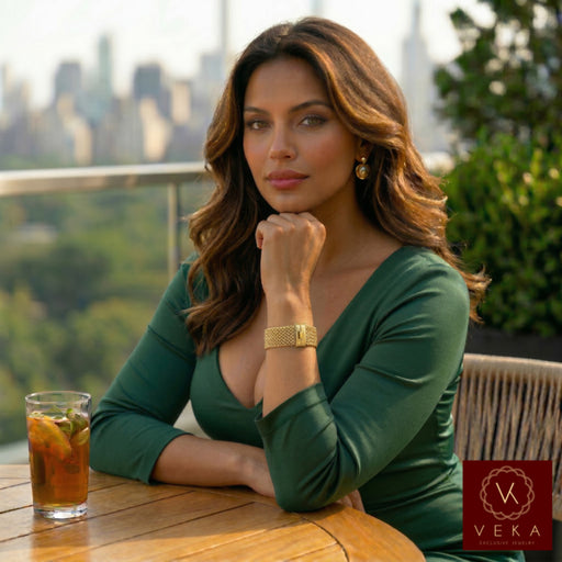 A high-resolution, professional photo of a model with long wavy brown hair, wearing a dark green V-neck dress. She is seated at a wooden table on a city terrace with a blurred skyline background. Her hand is resting under her chin, prominently displaying a thick, shimmering 14k gold popcorn-link bracelet on her wrist. The lighting is warm and natural, highlighting the detailed texture of the jewelry.