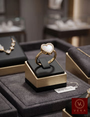 Close-up detail of a 14K yellow gold heart-shaped ring with an iridescent mother of pearl inlay and shimmering pavé diamond border, displayed on a finger in front of a grey velvet jewelry box