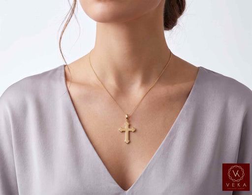 Woman wearing an authentic 14K yellow gold budded cross pendant on a delicate chain against a soft grey background
