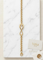 A high-resolution product flat-lay photograph of the Veka Jewelry 14K Yellow Gold Rolo chain bracelet. The bracelet, featuring its central infinity symbol charm with a clear CZ, is displayed full-length on a white Carrara marble slab. A branded Veka Jewelry linen business card is also visible on the marble