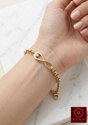 A close-up photograph of a woman’s wrist wearing the Veka Jewelry 14K Yellow Gold Rolo Link bracelet. The central polished gold infinity symbol charm features a single bezel-set clear cubic zirconia. Her hand is relaxed on a white marble surface, showing the delicate details of the semi-solid gold chain.
