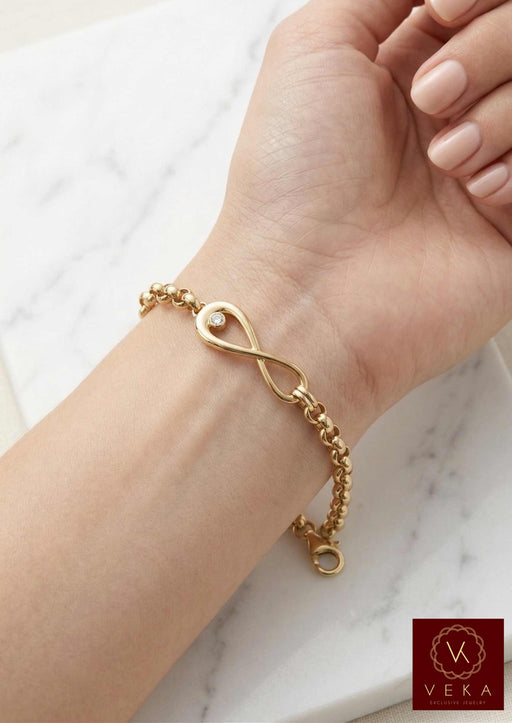 A close-up photograph of a woman’s wrist wearing the Veka Jewelry 14K Yellow Gold Rolo Link bracelet. The central polished gold infinity symbol charm features a single bezel-set clear cubic zirconia. Her hand is relaxed on a white marble surface, showing the delicate details of the semi-solid gold chain.