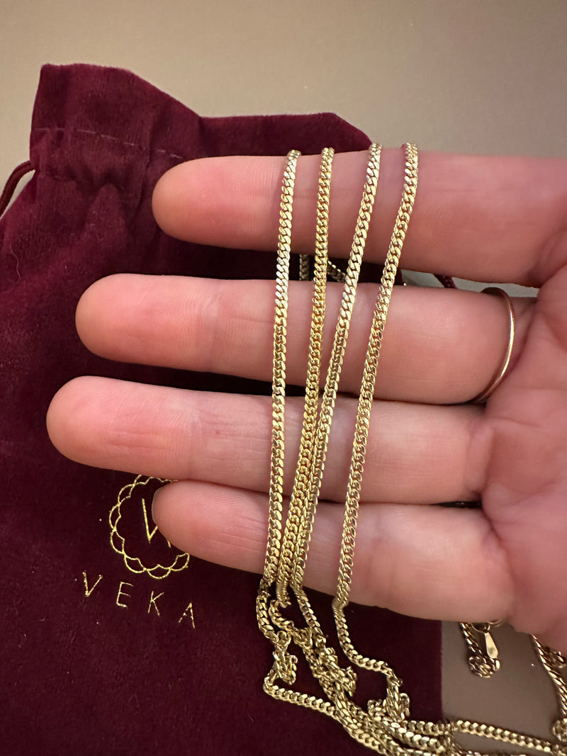Gold chains held by a hand with a maroon pouch labeled 'VEKA' in the background.