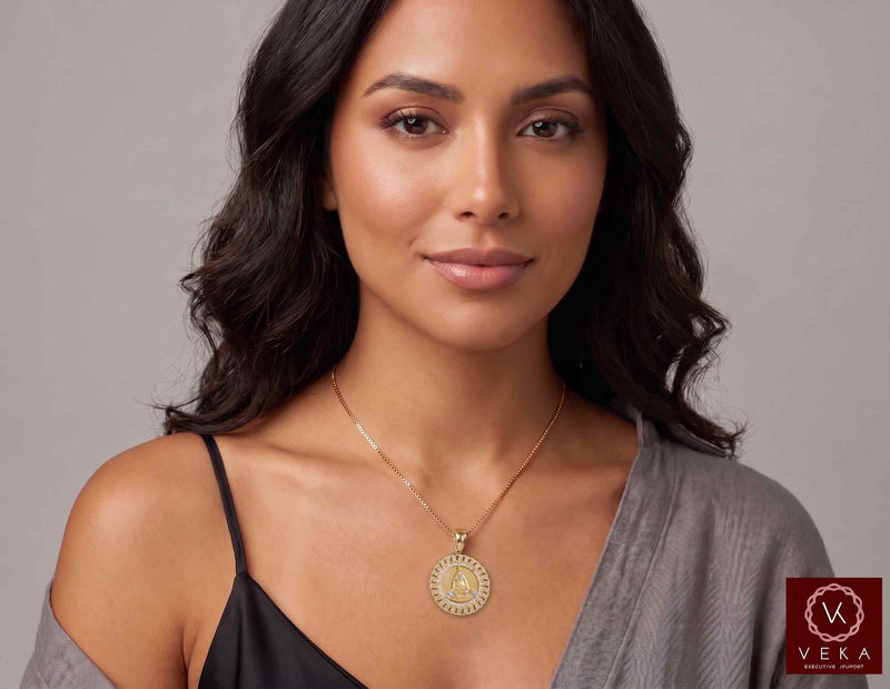 A woman with dark wavy hair models a gold Veka brand 14K Yellow Gold Cuban Link Border Caridad del Cobre Medal Pendant with CZ Accents with necklace, wearing a black camisole and a gray cardigan against a neutral gray background.