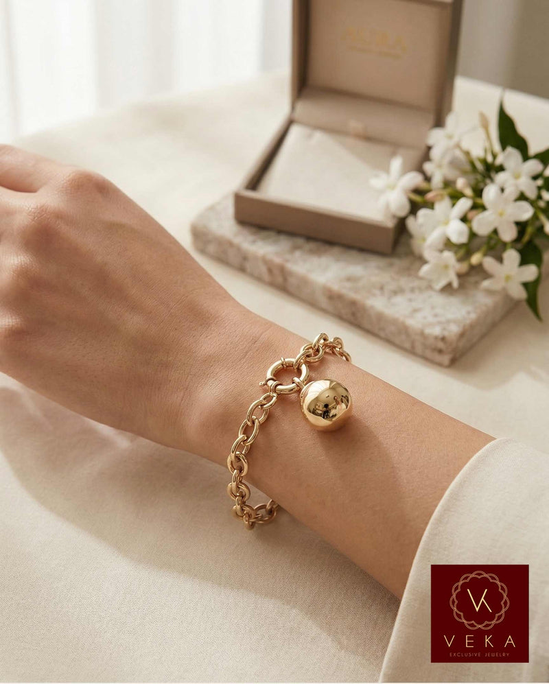 Lifestyle view of a woman wearing a 14K Yellow Gold Oval Rolo Link Bracelet with a polished sphere charm, styled with a luxury jewelry box and white flowers