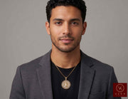 A man wearing a1 4K Yellow Gold Cuban Link Border Caridad del Cobre Medal Pendant with CZ Accents (9.1g) and necklace with a circular medallion featuring a religious figure, styled with a black t-shirt and a gray blazer against a neutral gray background. A Veka Jewelry brand logo is visible in the bottom right corner.