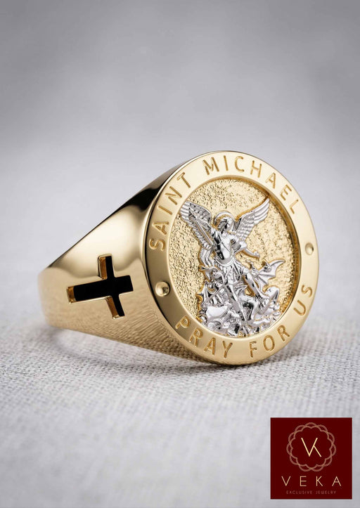 A detailed close-up product photo of a Men's 10K Two-Tone Gold Saint Michael Archangel Signet Ring, featuring a yellow gold band with a black side cross cutout and a highly detailed white gold depiction of St. Michael defeating the devil on a textured central face, with 'SAINT MICHAEL' and 'PRAY FOR US' embossed text.