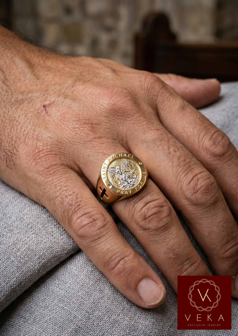 A photo showing a man's hand on a grey cloth surface, wearing the Men's 10K Two-Tone Gold Saint Michael Archangel Signet Ring on the ring finger, highlighting its size, fit, and design details on the hand.