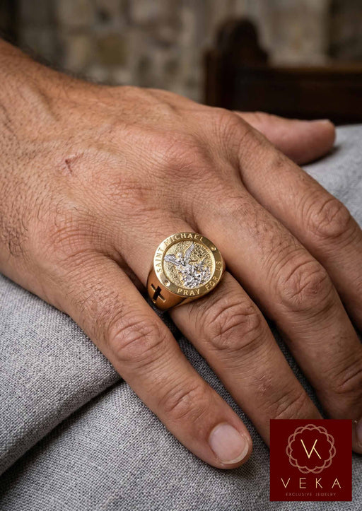 A photo showing a man's hand on a grey cloth surface, wearing the Men's 10K Two-Tone Gold Saint Michael Archangel Signet Ring on the ring finger, highlighting its size, fit, and design details on the hand.