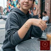 A teenage girl wearing a grey hoodie sits at an outdoor cafe table, showcasing a 3.5mm gold Cuban link bracelet with a cubic-zirconium-encrusted teddy bear charm featuring a synthetic ruby-red heart. She is also wearing thin gold stacking rings while a glass of iced coffee sits on the metal mesh table