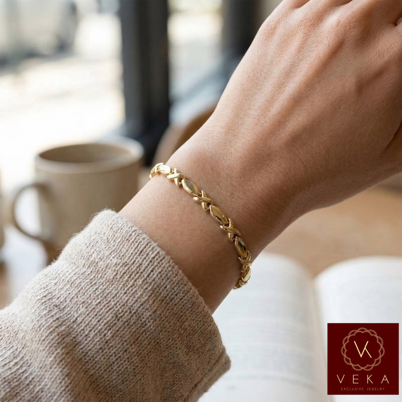 Close-up of a woman’s wrist wearing a thin 6mm 10K yellow gold XOXO bracelet in a casual cafe setting with a book.