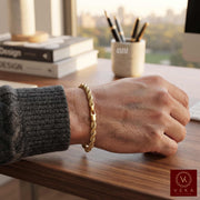 A 14k 4mm real gold rope-style chain bracelet worn on a person's wrist, resting on a wooden desk in a bright office setting. The person is wearing a dark gray ribbed sweater sleeve by Veka Jewelry