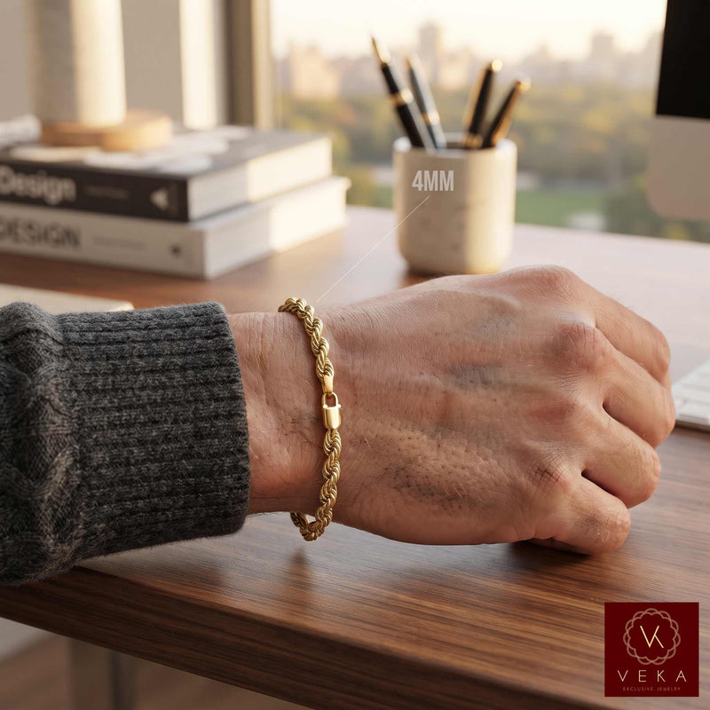 A 14k 4mm real gold rope-style chain bracelet worn on a person's wrist, resting on a wooden desk in a bright office setting. The person is wearing a dark gray ribbed sweater sleeve by Veka Jewelry