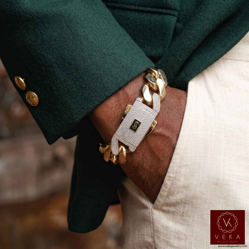 A close-up of a person's wrist wearing a Monaco 10K Real gold chain bracelet with a textured white a Pave Lock with The Monaco logo. The person is wearing a dark green blazer and off-white trousers, with a Veka brand watermark in the bottom right corner.