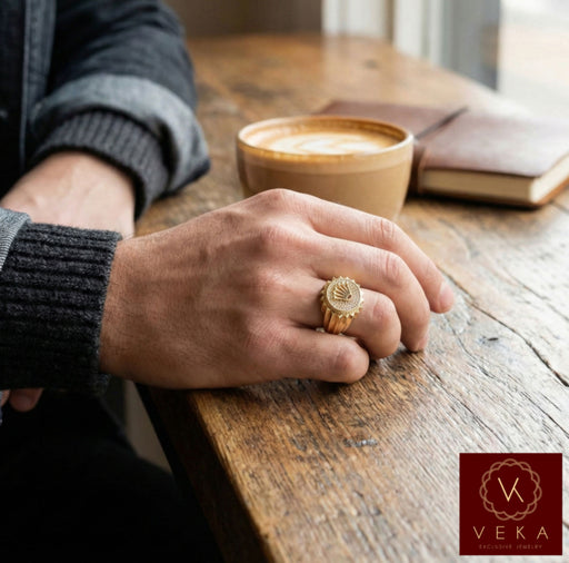 A close-up photograph of a man's hand wearing the Veka Jewelry 14K yellow gold crown signet ring with pave diamonds. His hand is resting on a wooden table next to a latte, showing a modern casual style with a denim jacket and knit sweater.