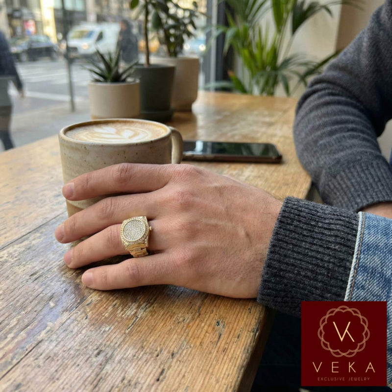 A close-up of a man's hand wearing the heavy Veka Jewelry 14K yellow gold watch-style ring with pave CZ stones. He is holding a ceramic coffee mug on a rustic wooden table in a modern cafe setting