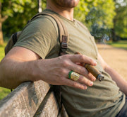 A close-up of a stylish man's hand holding a cigar, wearing the heavy Veka Jewelry 14K yellow gold Indian Chief ring. The ring features striking emerald green synthetic stones on the headdress and detailed gold craftsmanship.