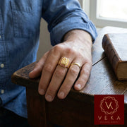 A stylish man wearing the Veka Jewelry 14K yellow gold Saint Barbara ring. The ring features a detailed gold relief of the Catholic Virgin surrounded by red synthetic rubies, showcased in a modern, professional setting.