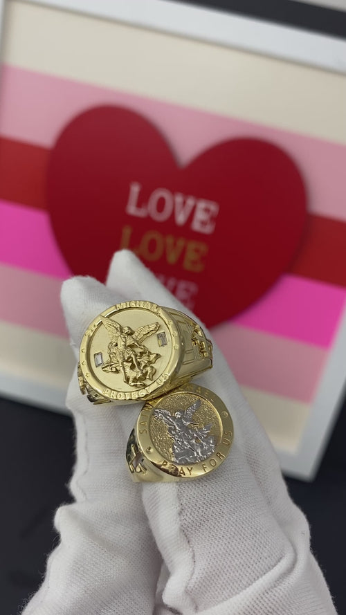 A close-up photograph of a white-gloved hand holding the 10K Two-Tone Gold Saint Michael Signet Ring, offering a focused view of the detailed central medal and text.