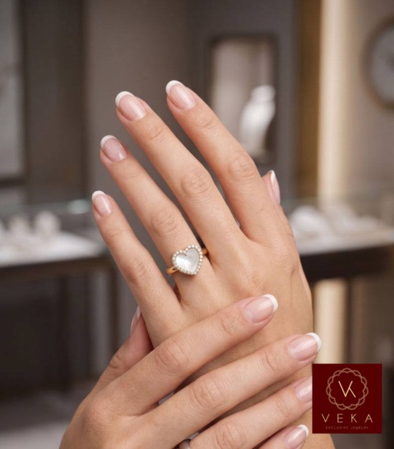 A hand model wearing the VEKA Exclusive Jewelry 14K yellow gold heart ring featuring a mother of pearl center surrounded by a diamond halo, styled with a classic French manicure in a luxury jewelry boutique setting