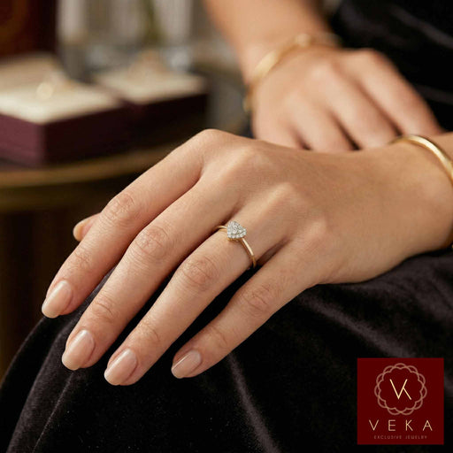 A model's hand with manicured nails wearing the delicate VEKA 10K yellow gold heart diamond cluster promise ring. Other blurred jewelry boxes are visible in the elegant background