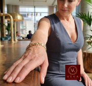 A woman wearing a Veka Jewelry 14K yellow gold heart and X link bracelet while wearing a modern grey V-neck dress in a luxury setting.